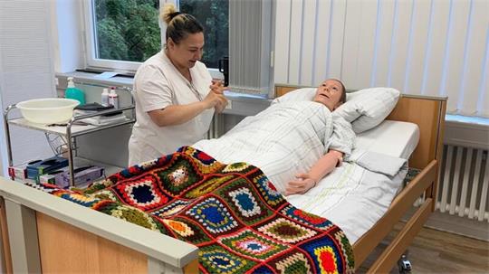 Eine Auszubildende in der Pflege steht an einer Pflegepuppe, die in einem Bett liegt.