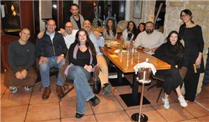 Gruppe von Menschen sitzt und steht um einen langen Holztisch in einem Restaurant mit Steinwand und Fliesenboden.