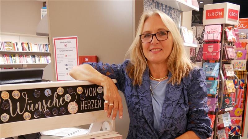 Frau mit blonden Haaren und blauem Blazer lehnt an einem Kassenbereich mit der Aufschrift 'Wünsche von Herzen' in einer Buchhandlung