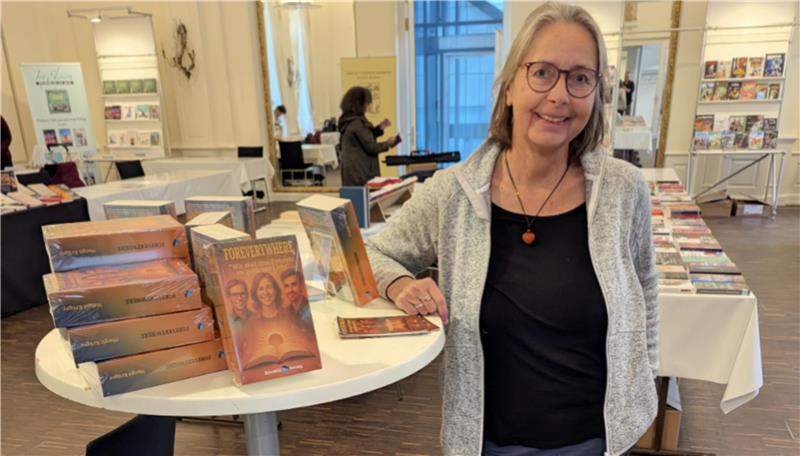 Frau mit grauem Cardigan und schwarzem Shirt steht an rundem Tisch mit mehreren Stapeln von Büchern in einem hellen Raum mit Bücherregalen.