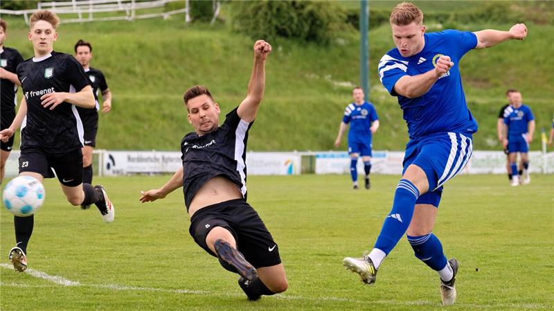 Fußballspieler in blauer und schwarzer Sportkleidung kämpfen auf einem Spielfeld um den Ball, ein Spieler in Schwarz rutscht auf dem Boden liegend zur Abwehr