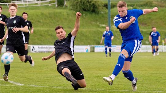 Fußballspieler in blauer und schwarzer Sportkleidung kämpfen auf einem Spielfeld um den Ball, ein Spieler in Schwarz rutscht auf dem Boden liegend zur Abwehr