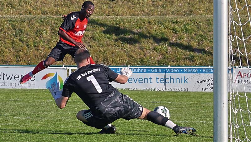 Fußballspieler in rotem Trikot schießt Ball am Torwart in schwarzem Trikot vorbei auf das Tor
