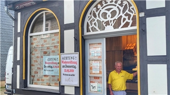 Fassade eines Fachwerkhauses mit zwei großen Fenstern und einer Tür, davor steht ein Mann in gelbem Hemd