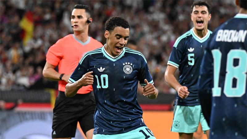 Ein Fußballspieler in einem blauen Trikot jubelt über sein Tor, links hinter ihm ein Schiedsrichter, der ein rotes Trikot trägt, am Bildrand zwei weitere Fußballer in blauen Trikots.