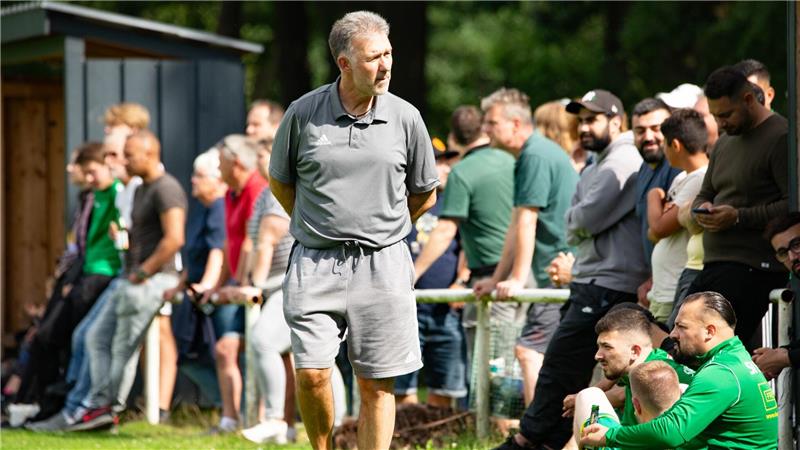 Mann in grauem T-Shirt und Shorts steht auf grünem Rasen vor einer Gruppe von Menschen, die an einem Geländer lehnen oder sitzen