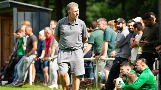 Mann in grauem T-Shirt und Shorts steht auf grünem Rasen vor einer Gruppe von Menschen, die an einem Geländer lehnen oder sitzen