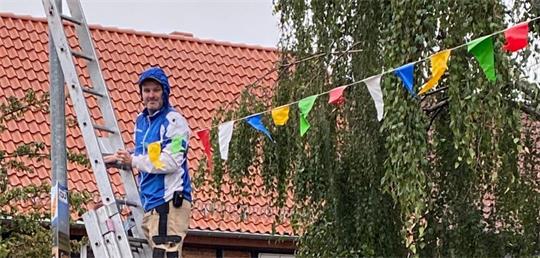 Mann in blauer und weißer Jacke steht auf einer Leiter vor einem Haus mit rotem Ziegeldach und bunten Wimpeln im Hintergrund