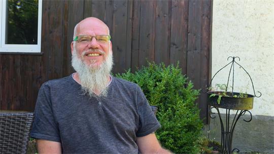 Mann mit Brille und langem Bart sitzt lächelnd vor einer Holzfassade neben einem Busch