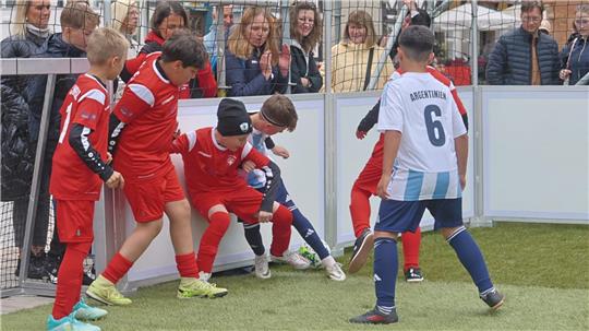 Mehrere Kinder in Fußballtrikots kämpfen um den Ball nahe einer Bande auf einem Kunstrasenplatz