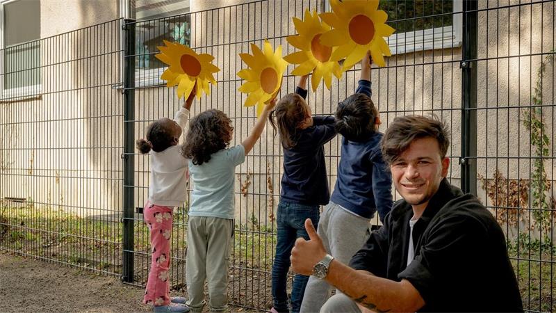 Vier Kinder befestigen große gelbe Sonnenblumen an einem neuen Metallzaun. Daneben kniet ein Mann und zeigt mit erhobenem Daumen in die Kamera.