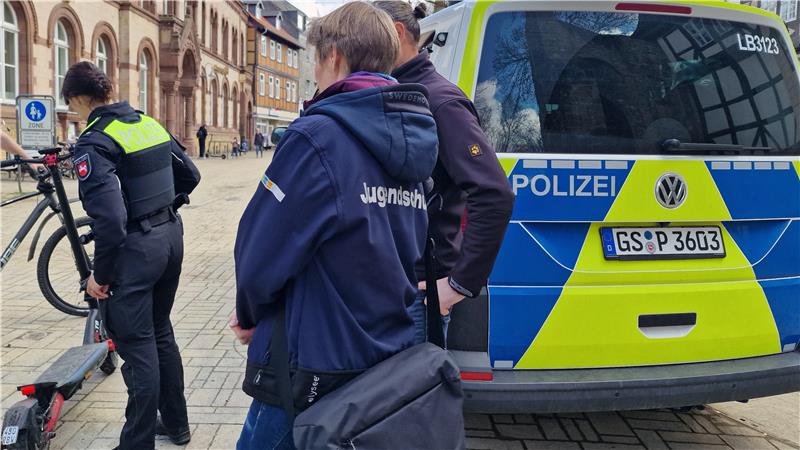 Polizeiwagen mit blau-gelber Folierung und drei Personen in Uniform auf einem gepflasterten Platz vor einem historischen Gebäude.