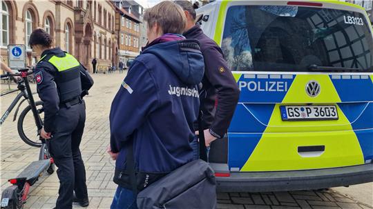 Polizeiwagen mit blau-gelber Folierung und drei Personen in Uniform auf einem gepflasterten Platz vor einem historischen Gebäude.