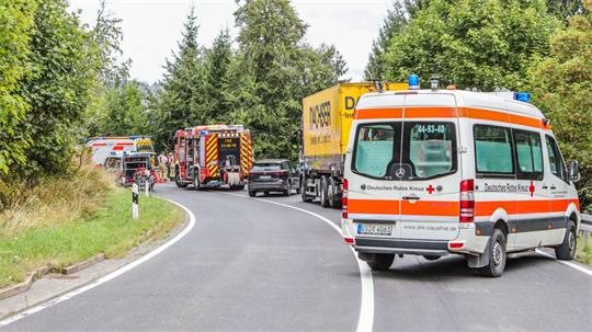 Mehrere Einsatzfahrzeuge, darunter ein Rettungswagen und Feuerwehrfahrzeuge, stehen auf einer kurvigen Landstraße in einer bewaldeten Gegend