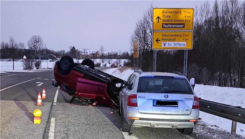 Zwei Autos auf einer schneebedeckten Landstraße, eines liegt auf dem Dach, das andere steht daneben, im Hintergrund gelbe Verkehrsschilder mit Ortsangaben