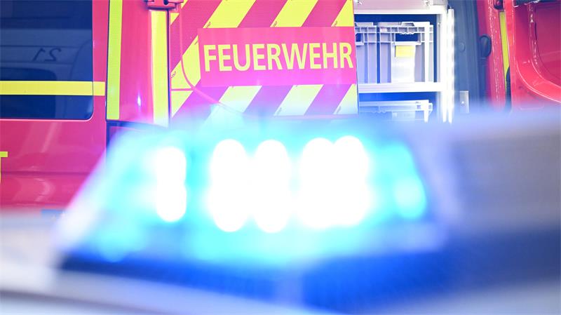 Ein Fahrzeug der Feuerwehr steht bei einem Einsatz auf einer Straße, während im Vordergrund das Blaulicht eines Polizeifahrzeugs zu sehen ist. 