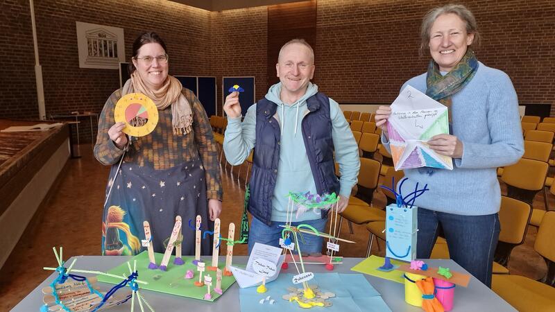 Drei Personen stehen hinter einem Tisch mit bunten Bastelarbeiten und halten selbstgebastelte Objekte in der Hand, im Hintergrund Reihen von Stühlen und eine Wand mit Holzvertäfelung.