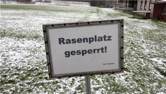 Schild mit der Aufschrift 'Rasenplatz gesperrt!' vor einem schneebedeckten Rasenplatz und Wald im Hintergrund.