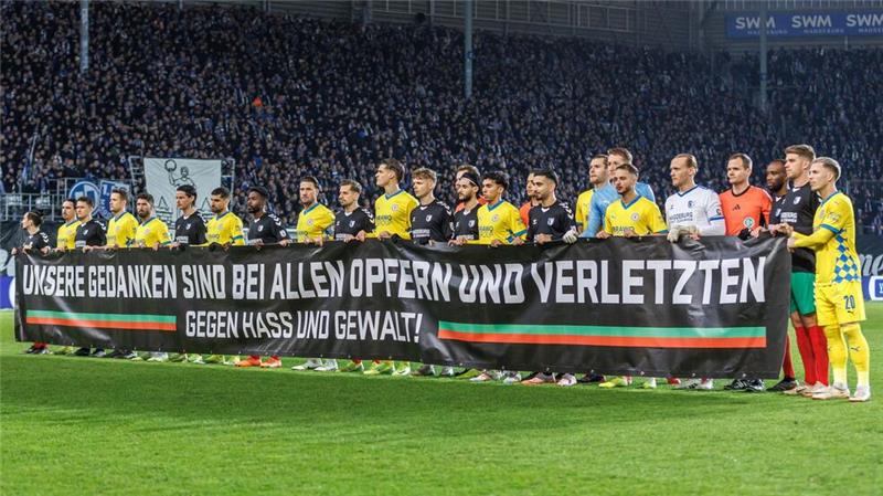 Beide Teams stehen während einer Schweigeminute zusammen hinter einem Transparent mit der Aufschrift: „Unsere Gedanken sind mit allen Opfern und Verletzten - gegen Hass und Gewalt!“.