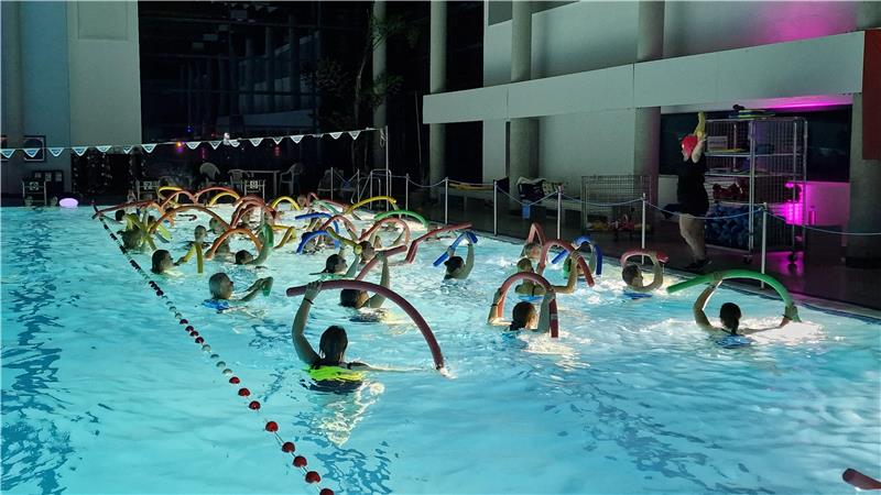 Gruppe von Menschen im Schwimmbad, die mit Schwimmnudeln im Wasser stehen und einer Person an der Beckenwand zuhören.