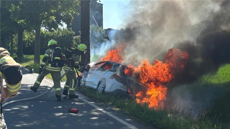 Riesige Flamen schlagen aus dem Auto, Feuerwehrmänner richten einen Schlauch mit Wasser auf das Auto