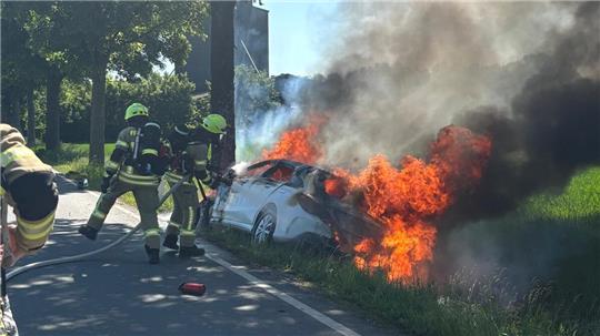 Riesige Flamen schlagen aus dem Auto, Feuerwehrmänner richten einen Schlauch mit Wasser auf das Auto