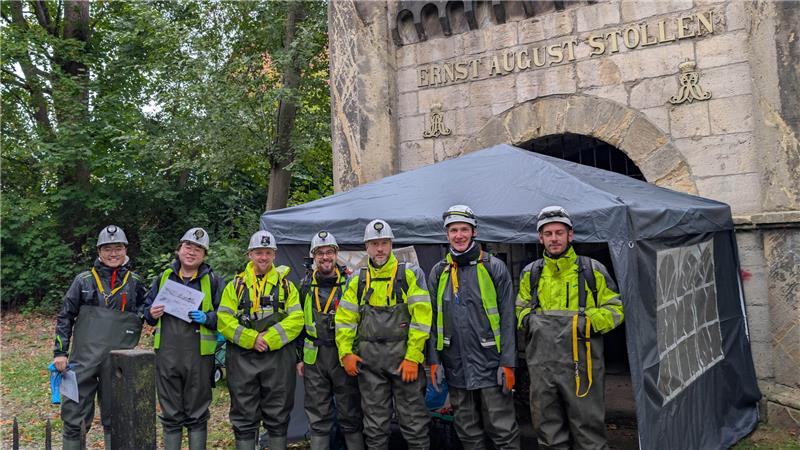 Sieben Personen mit Latzhosen, Gummistiefeln und Helmen stehen in einer Reihe vor dem Eingang zum Ernst-August Stollen.