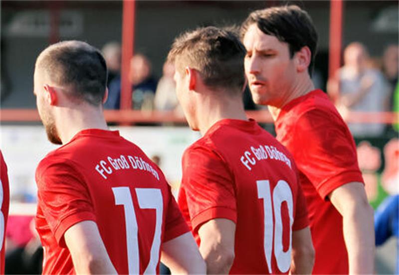 Drei Fußballspieler in roten Trikots stehen nebeneinander vor einem Schiedsrichter auf dem Spielfeld.