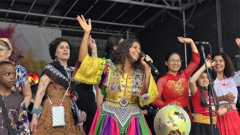 Mehrere Menschen auf einer Bühne, eine Frau in bunter traditioneller Kleidung singt mit Mikrofon und hebt die Hand.