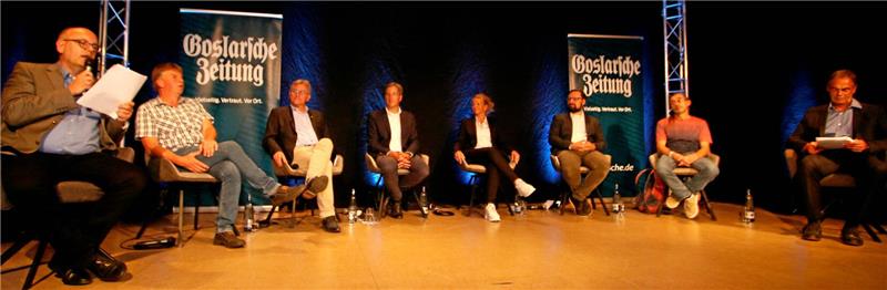 Acht Personen sitzen auf Stühlen auf einer Bühne mit zwei Bannern der "Goslarschen Zeitung" im Hintergrund.