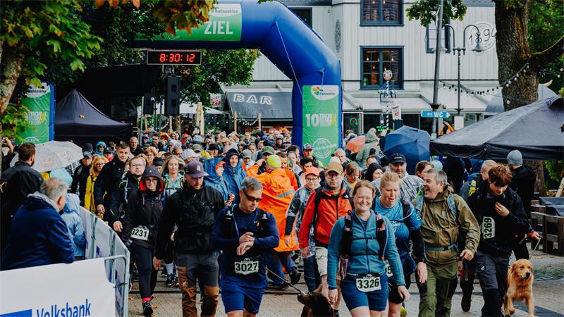 Menschenmenge bei einem Wandermarathon mit bunten Jacken und Regenschutz vor einem blauen Zielbogen.