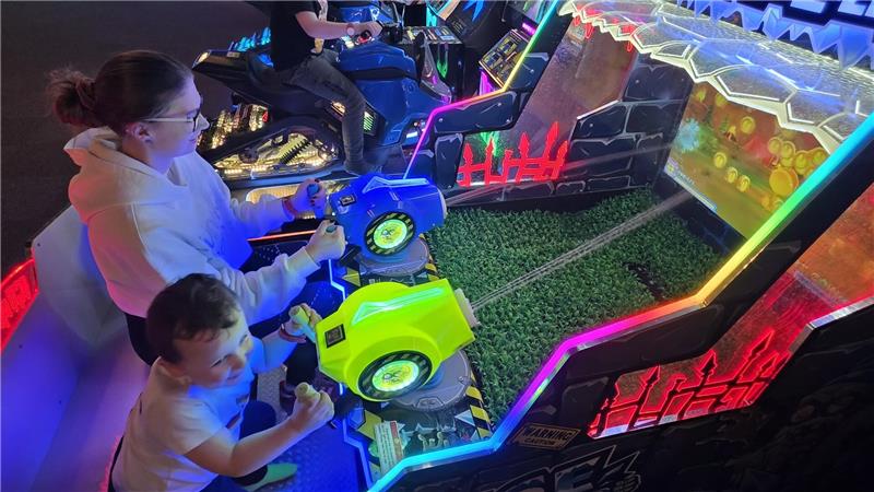 Zwei Personen sitzen an einem leuchtenden Arcade-Spiel mit Motorradlenkern und einem Spielfeld mit grüner Oberfläche.