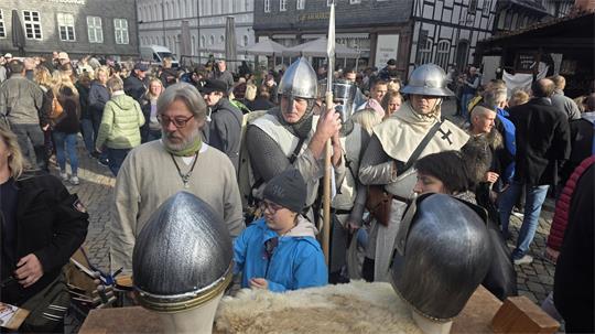 Menschenmenge auf einem Marktplatz mit zwei Männern in mittelalterlicher Rüstung und Helmen auf einem Tisch im Vordergrund.