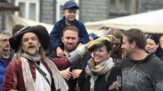 Mann in historischer Kleidung legt einem lächelnden Mädchen eine Mütze auf den Kopf, umgeben von Erwachsenen und Kindern in moderner Kleidung auf einem Fest
