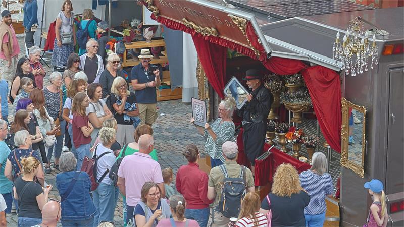Menschenmenge vor einem Straßenkünstler mit schwarzem Hut und schwarzem Mantel, der vor einem mit roten Vorhängen und goldenen Verzierungen geschmückten Stand auf Kopf steht.