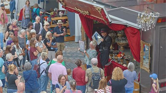 Menschenmenge vor einem Straßenkünstler mit schwarzem Hut und schwarzem Mantel, der vor einem mit roten Vorhängen und goldenen Verzierungen geschmückten Stand auf Kopf steht.