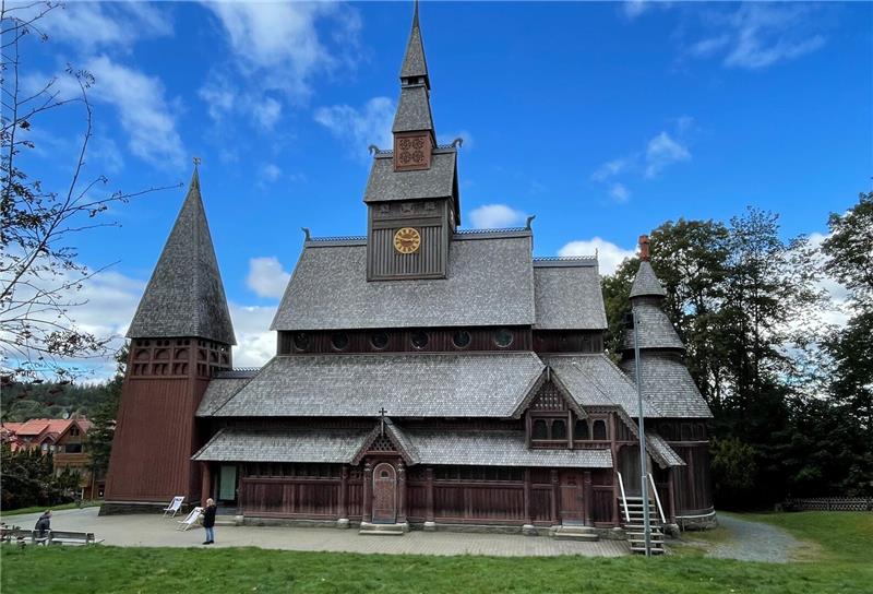 Märchentante, Hase und Förster geben sich die Klinke in die Hand Eine Holzstabkirche vor blauem Himmel auf grüner Wiese.