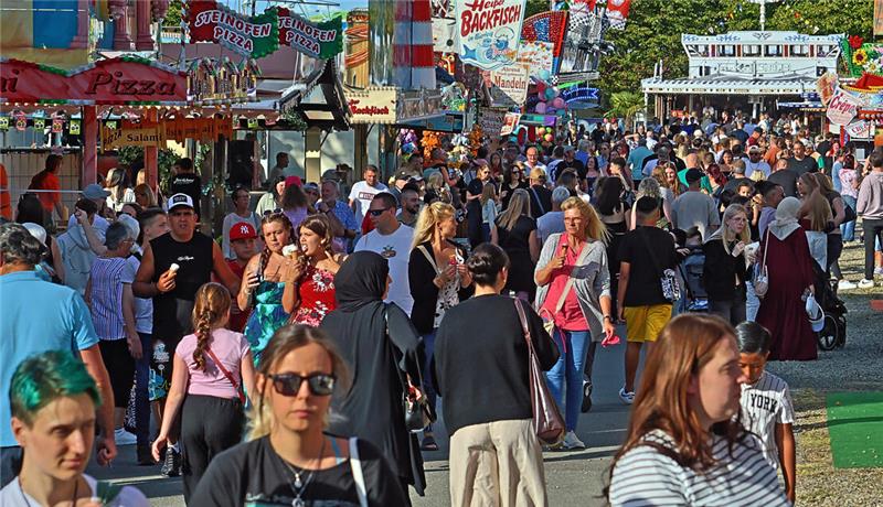 Menschenmenge auf einem belebten Festgelände mit bunten Ständen und Werbeschildern im Hintergrund.