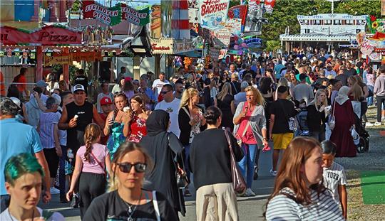 Menschenmenge auf einem belebten Festgelände mit bunten Ständen und Werbeschildern im Hintergrund.