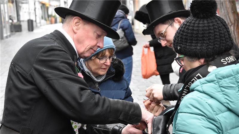 Der GZ-Schweinchenverkauf läuft in Goslar auf Hochtouren Zwei Männer in schwarzen Fräcken und Zylindern geben einem Kind in blauer Jacke und schwarzer Mütze etwas in die Hand auf einer Straße mit Passanten im Hintergrund
