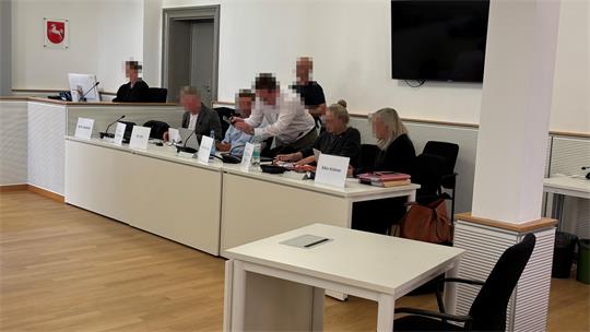 Mehrere Personen sitzen an einem langen Tisch in einem hellen Raum mit Holzfußboden und weißen Wänden