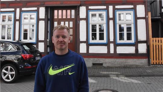 Ein Mann steht im blauen Pullover vor einem Haus.