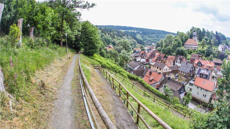 Wanderweg mit Holzzaun führt an bewaldetem Hang vorbei zu einem Dorf mit Fachwerkhäusern und Kirche im Hintergrund