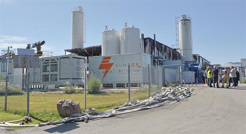 Industrieanlage mit mehreren hohen Silos und einem Zaun im Vordergrund, vor dem ein großer Stein und ein Bündel Kabel liegen, im Hintergrund mehrere Personen auf einem Weg