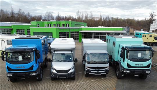 Vier Lastwagen verschiedener Modelle, zwei in Blau und zwei in Weiß, stehen nebeneinander auf einem gepflasterten Parkplatz vor einem grünen Gebäude.