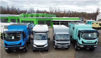 Vier Lastwagen verschiedener Modelle, zwei in Blau und zwei in Weiß, stehen nebeneinander auf einem gepflasterten Parkplatz vor einem grünen Gebäude.