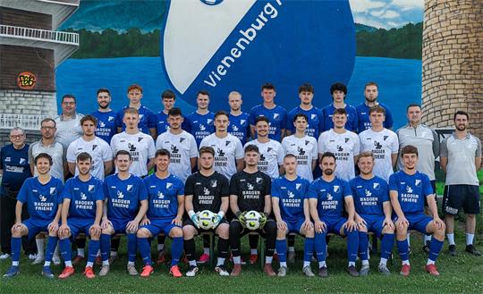Mannschaftsfoto einer Fußballmannschaft mit Spielern und Trainern vor einer Wand mit großem Vereinswappen und zwei Türmen