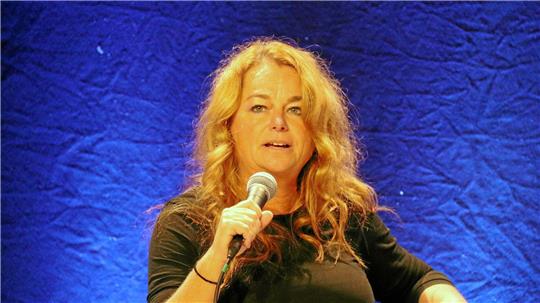 Frau mit langen, lockigen Haaren hält ein Mikrofon vor blauem Hintergrund.