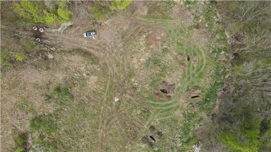 Luftaufnahme einer Wiese mit mehreren Erdhügeln, einem Auto und umgebenden Bäumen