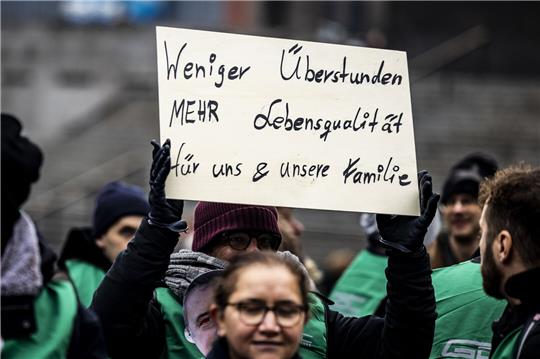 Eine Demonstrantin hält ein Plakat in die Höhe, sie wendet sich gegen Überstunden.